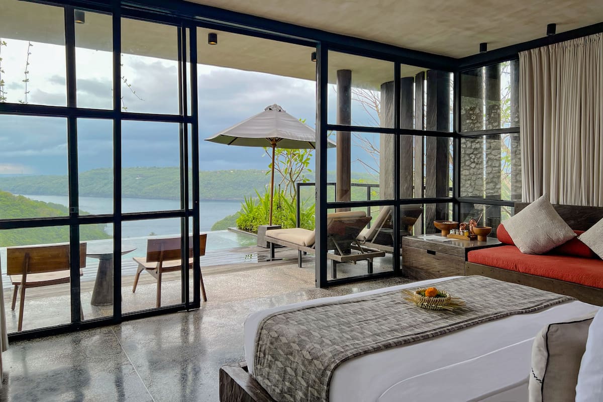 A spacious bedroom with a large bed, a seating area with a table and chairs, and a view of a lake through floor-to-ceiling windows.