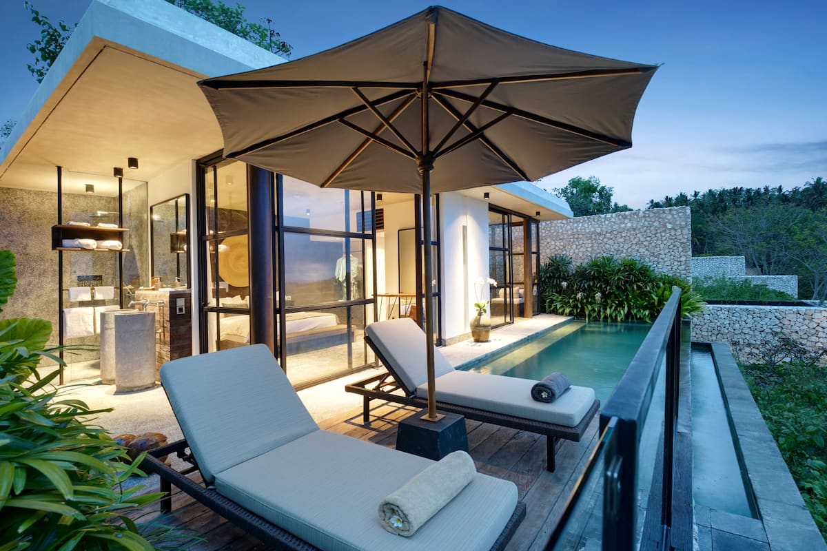 A modern outdoor pool area with lounge chairs and a large umbrella, surrounded by a stone wall and lush greenery.