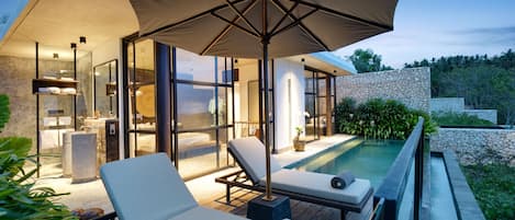 A modern outdoor pool area with lounge chairs and a large umbrella, surrounded by a stone wall and lush greenery.