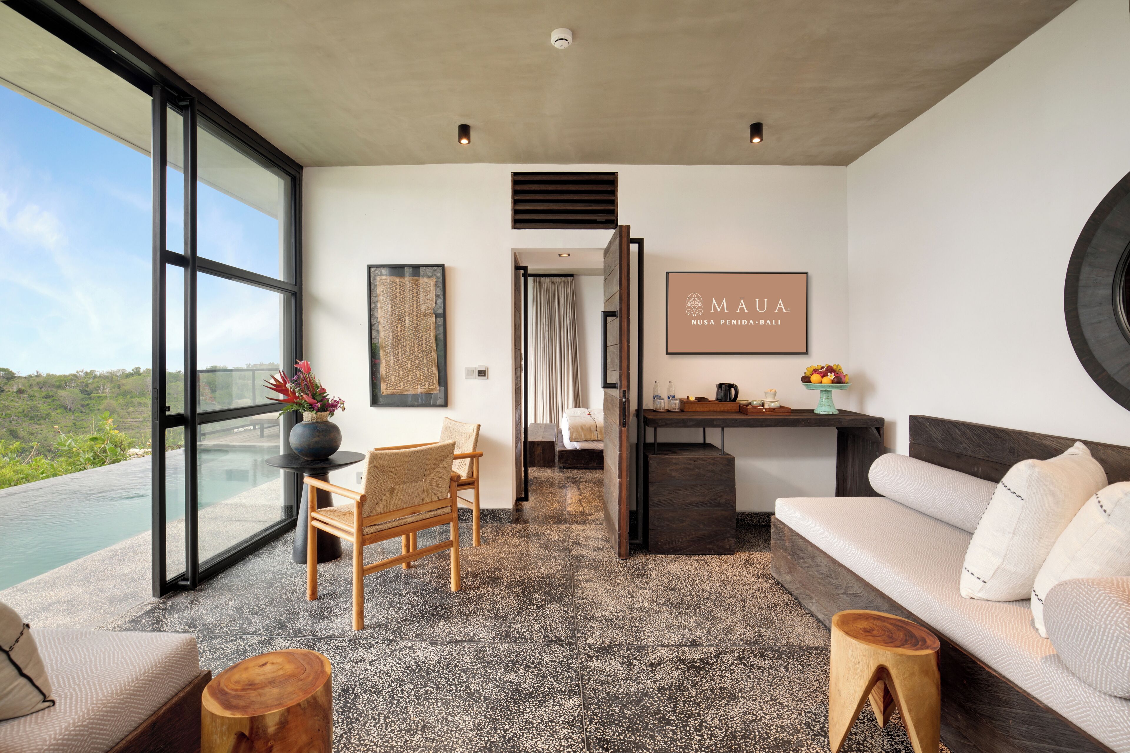 Una habitación de hotel moderna con un ventanal grande, un sofá, una silla de madera, una mesita y un televisor en la pared.