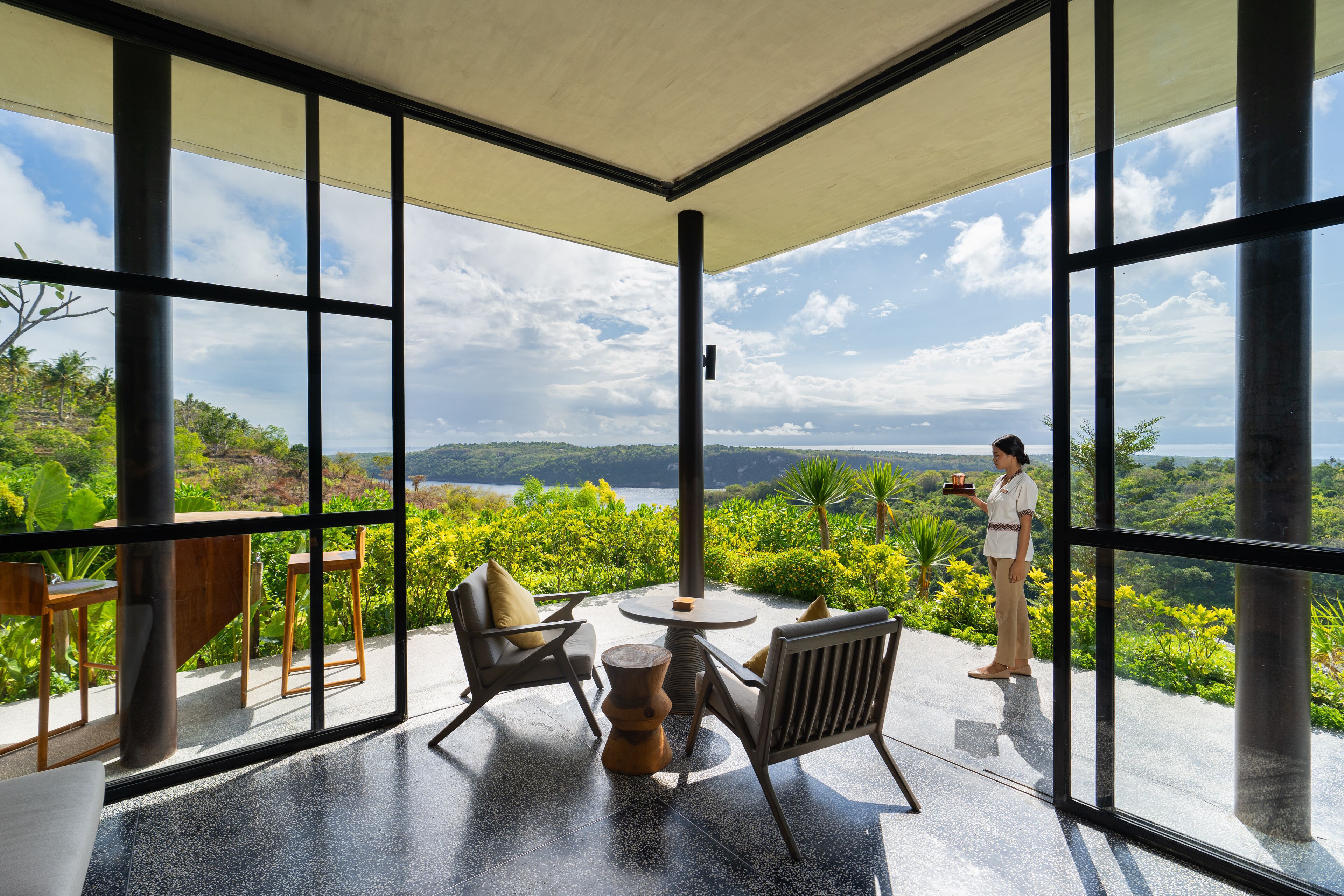 A spacious balcony with a view of a lake and greenery, furnished with chairs and a table.