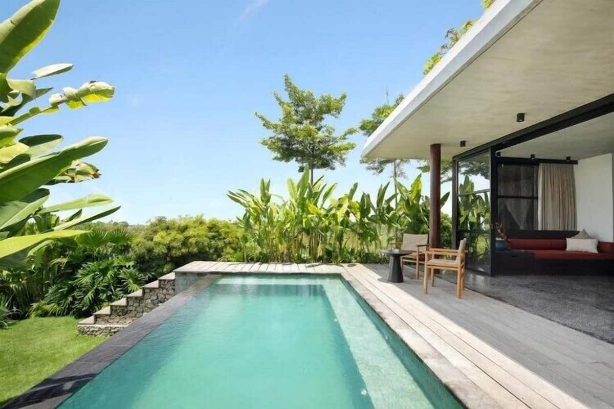 A modern outdoor pool area with a wooden deck, surrounded by lush greenery and a clear blue sky.