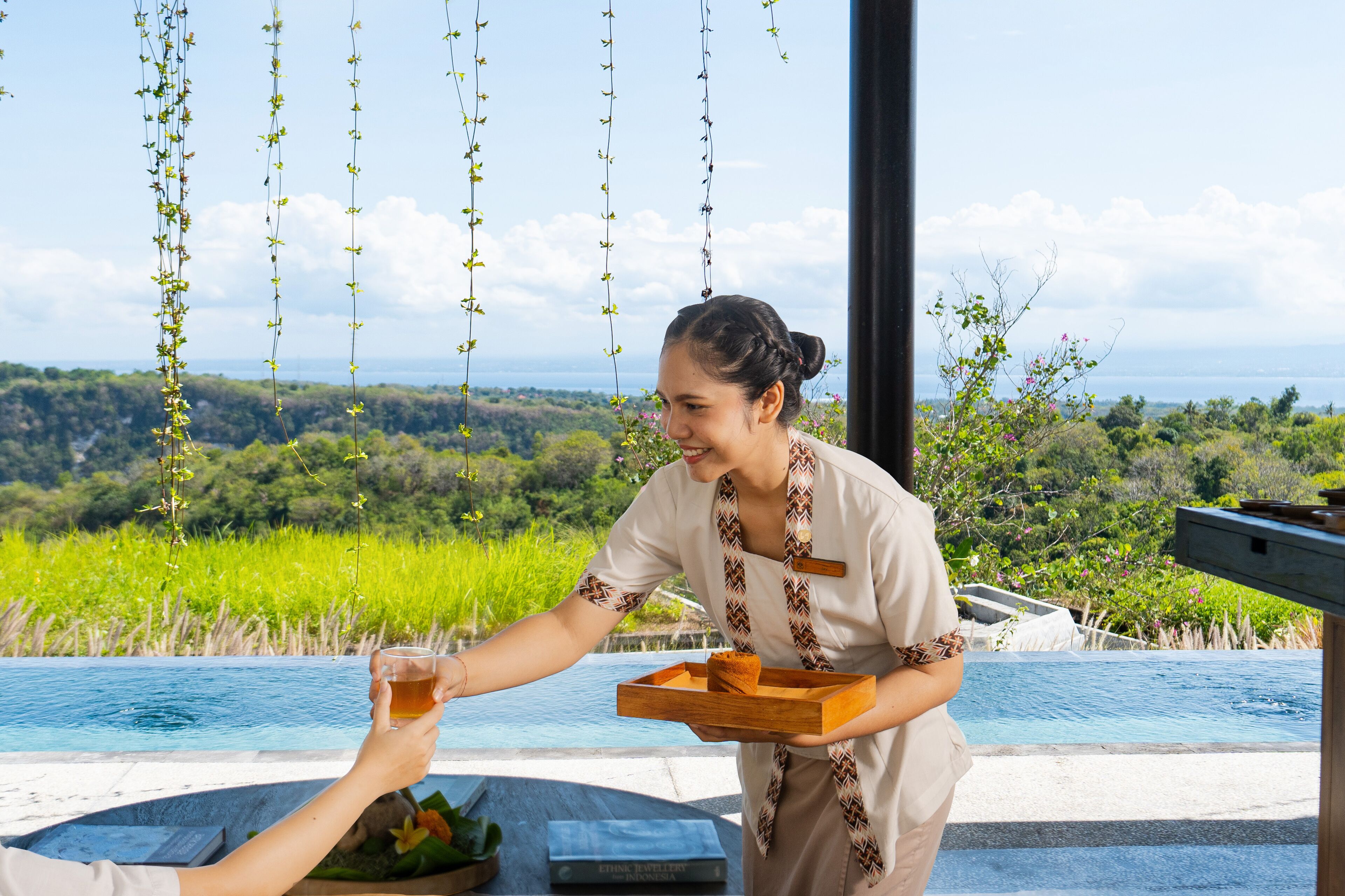 Une personne sert une boisson et apporte un plateau avec un dessert à une table située au bord de la piscine, offrant une vue panoramique.