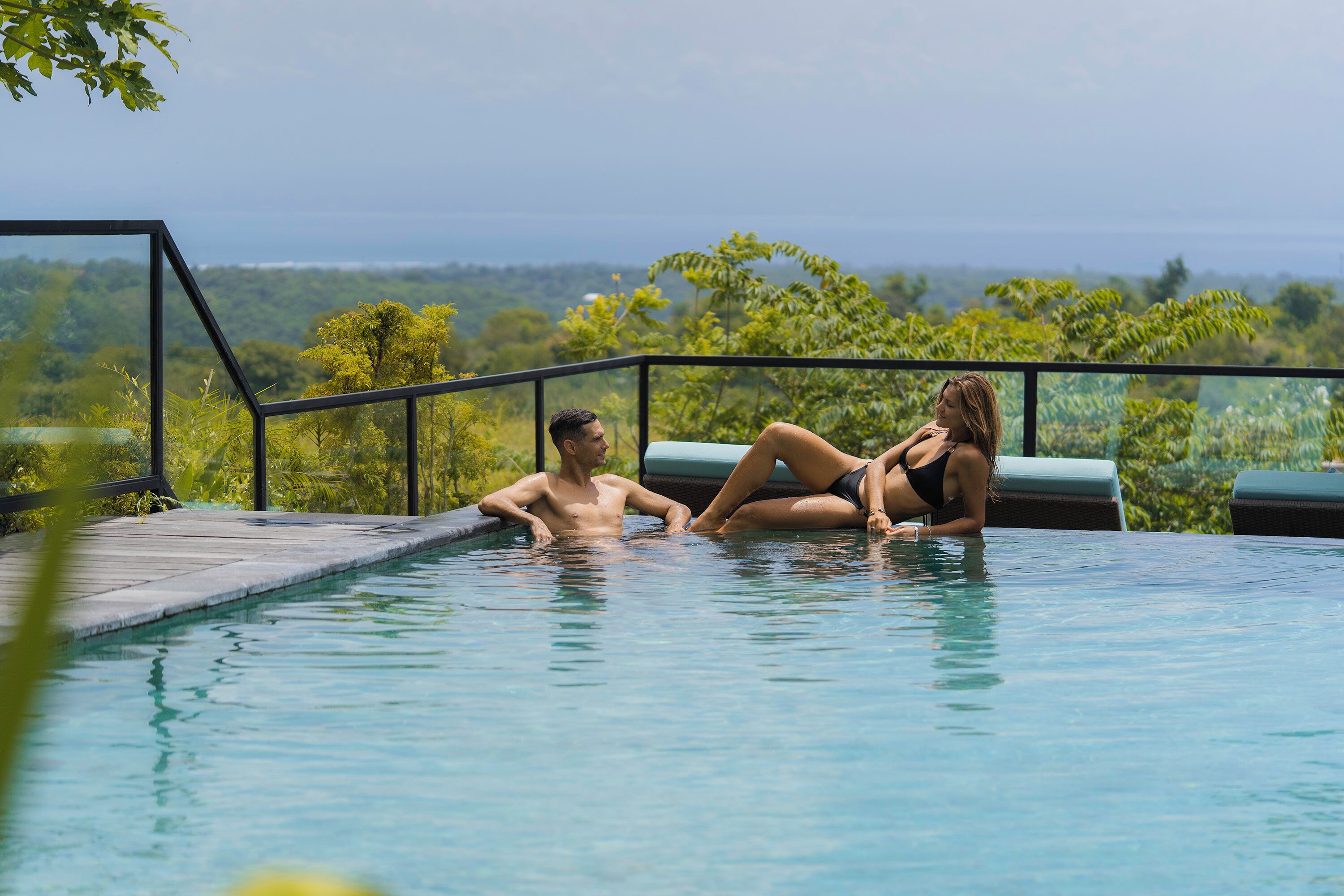 A couple relaxing in a pool with a view of the ocean and greenery.