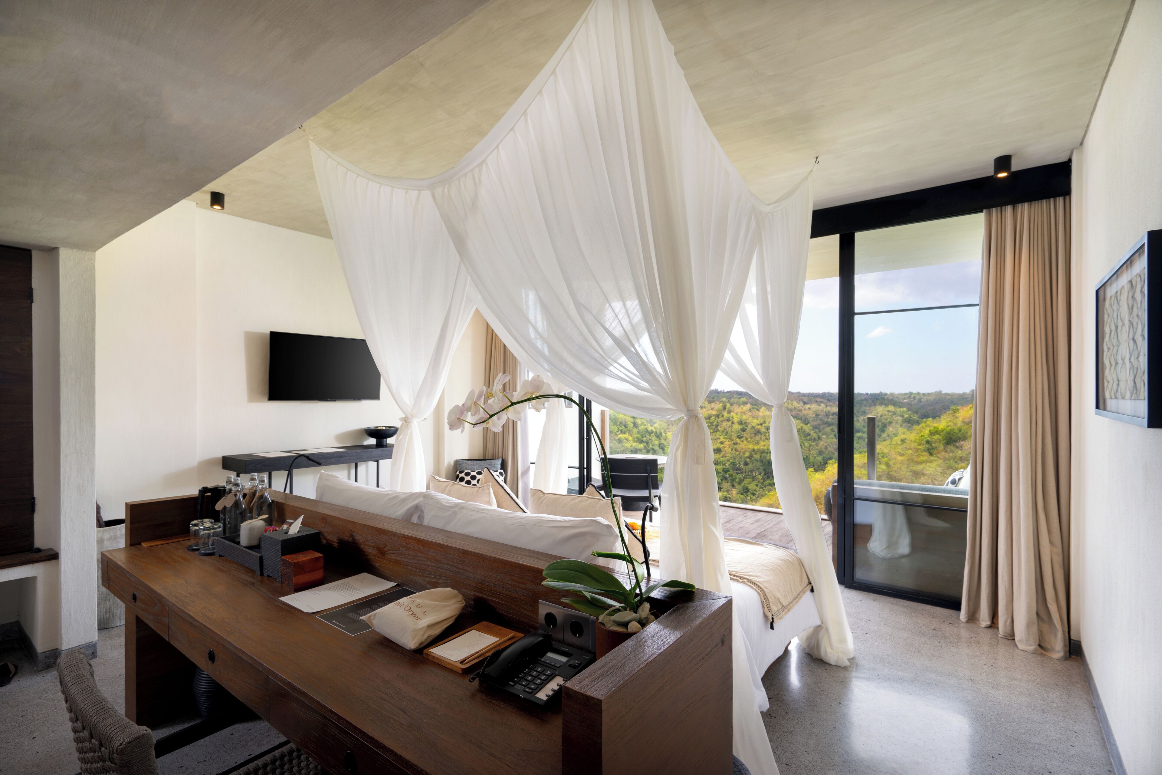 Une chambre moderne avec un bureau en bois, un lit à baldaquin, un téléviseur fixé au mur et un balcon offrant une vue sur le paysage.