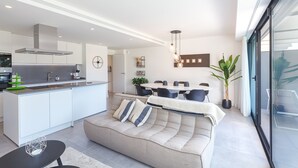 Una cocina moderna con una isla central, un comedor con una mesa redonda y sillas, un sofá con cojines y un ventanal con vistas.