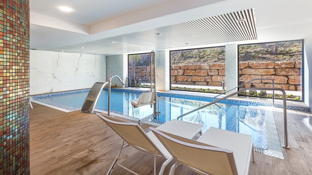 Una piscina interior moderna con paredes de vidrio transparente, suelo de madera y vista a un paisaje rocoso.