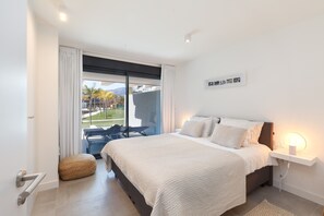 A bedroom with a large bed, a bedside table with a lamp, a view of the outdoors, and a small seating area.