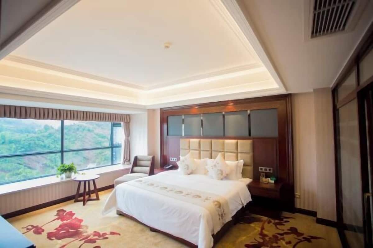 A hotel room with a large bed, a small table, a chair, a window with a view, and a ceiling light.