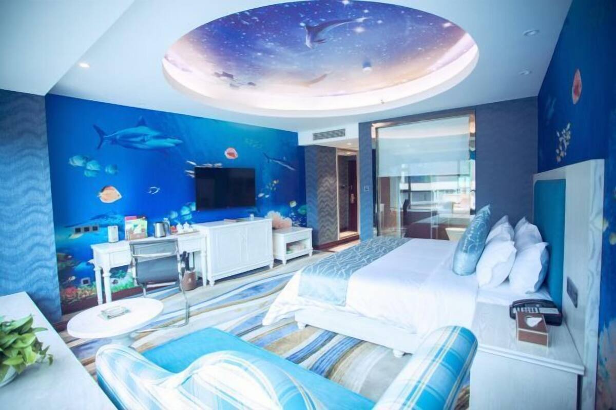 A bedroom with a bed, a desk, a chair, a TV, and a ceiling mural.