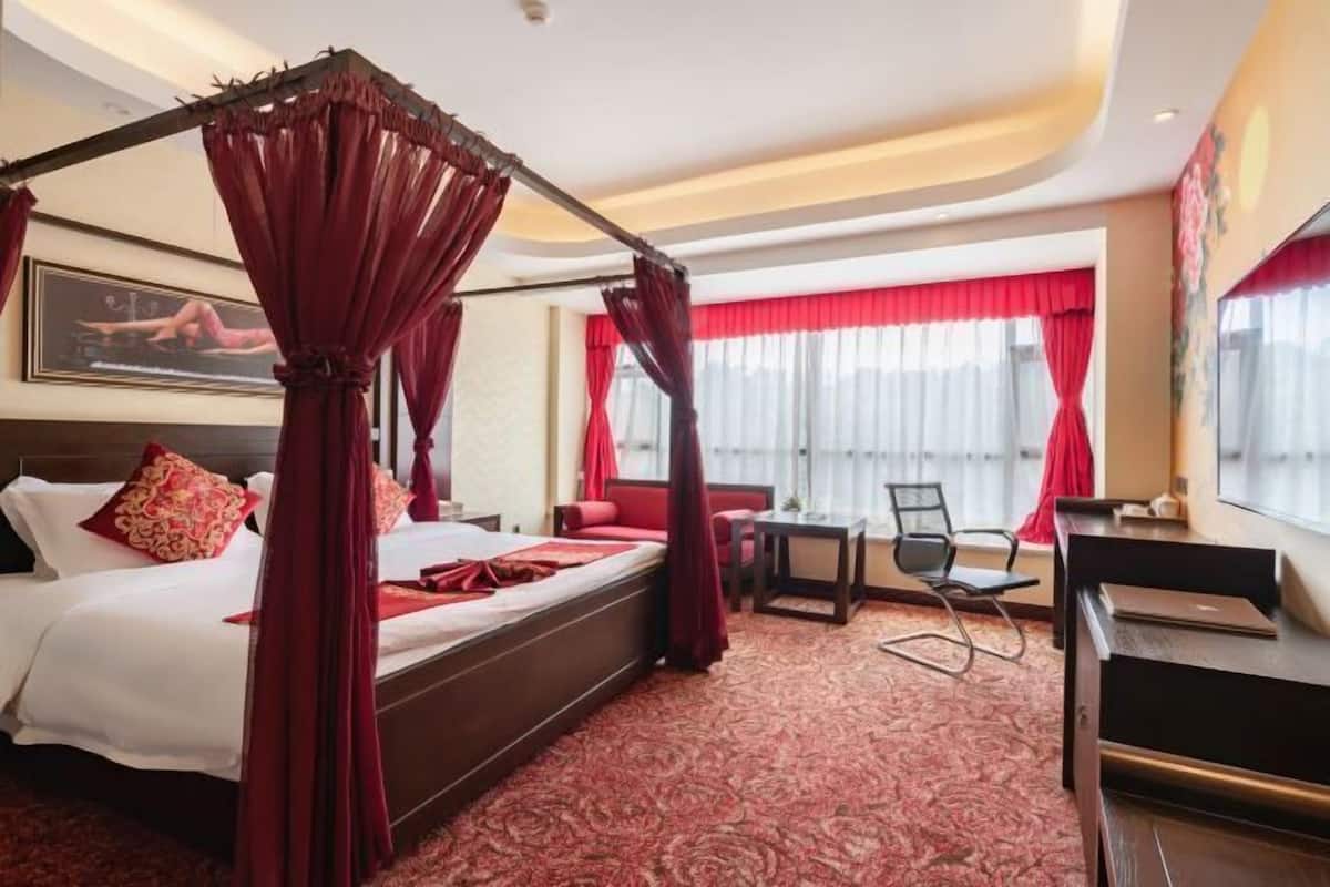 A hotel room with a four-poster bed, a desk, a chair, and a view of the city.