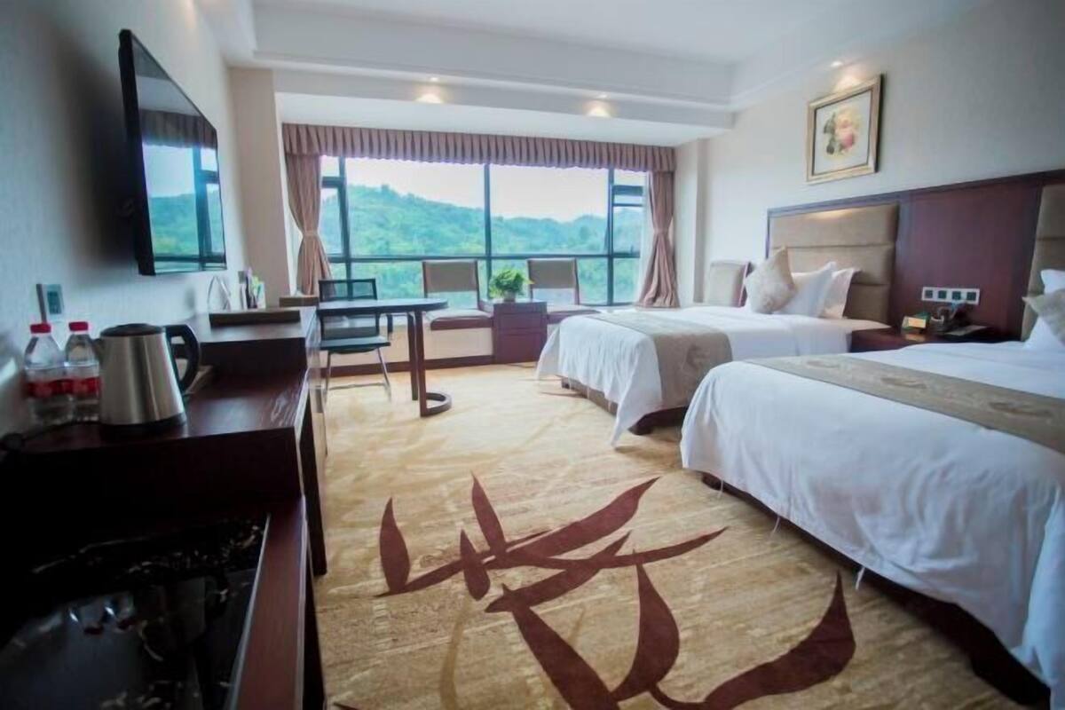 A hotel room with two beds, a desk with a chair, a TV, and a large window offering a view of greenery.
