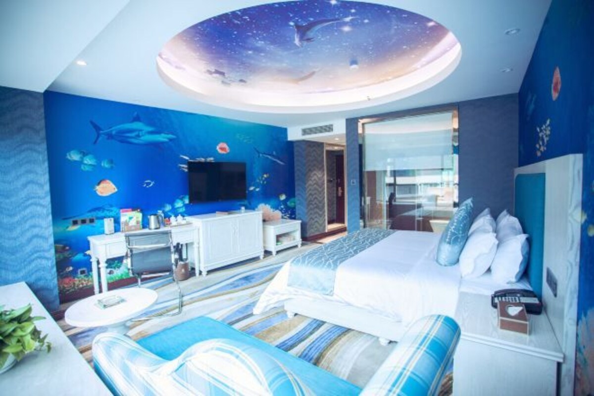 A bedroom with a bed, a desk, a chair, a TV, and a ceiling mural.