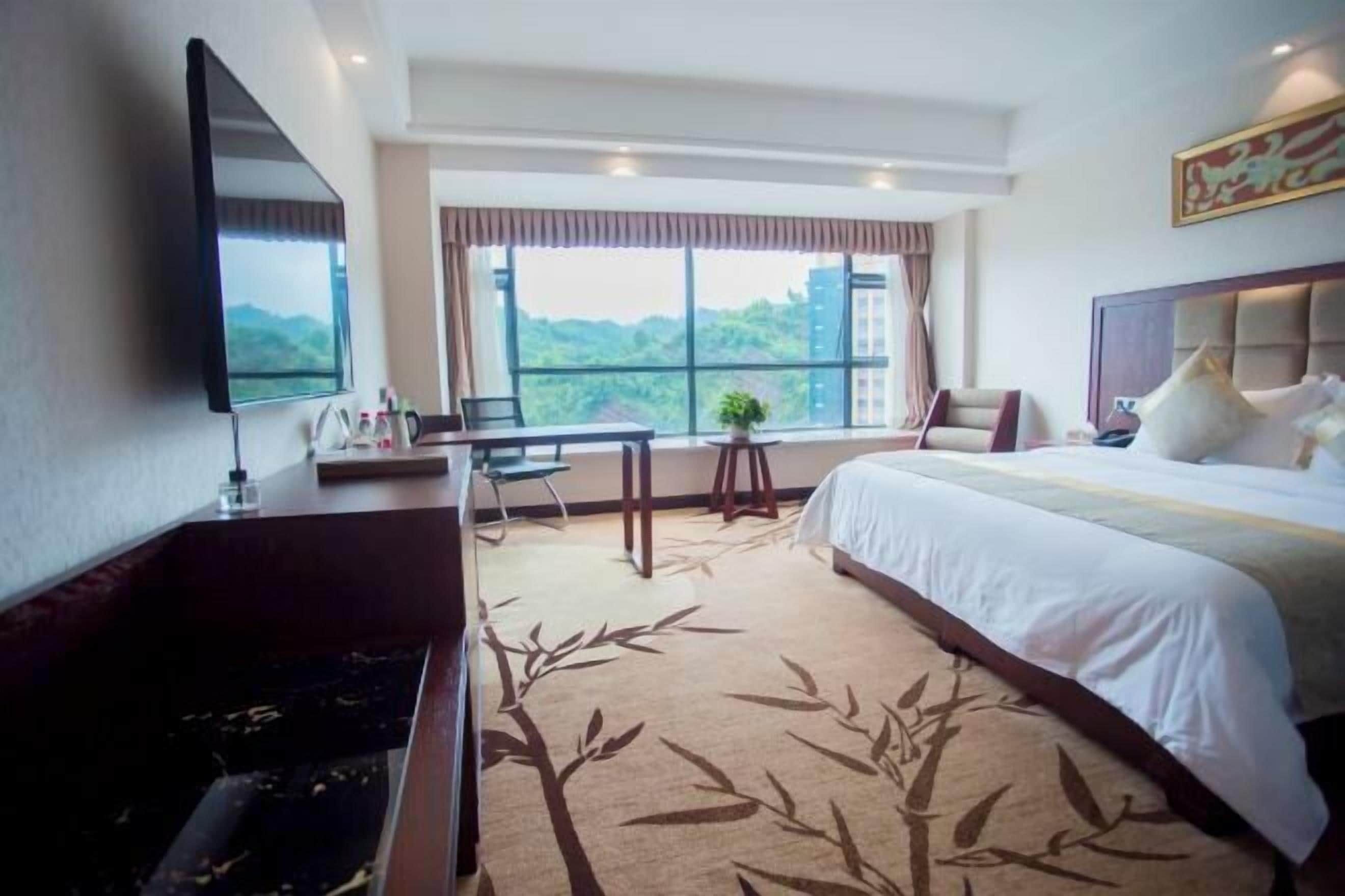 Habitación de hotel con cama, escritorio, silla, televisión y ventana grande con vistas a follaje.