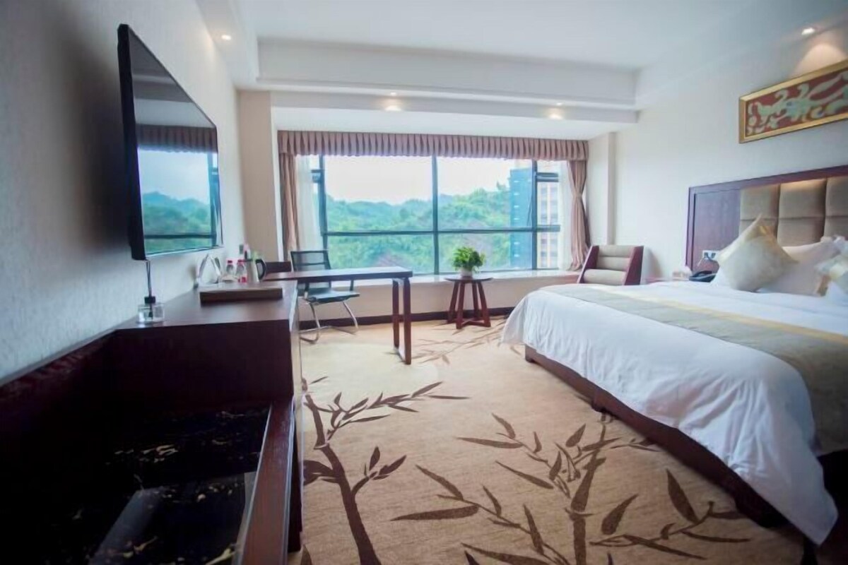 A hotel room with a bed, a desk, a chair, a TV, and a large window with a view of greenery.
