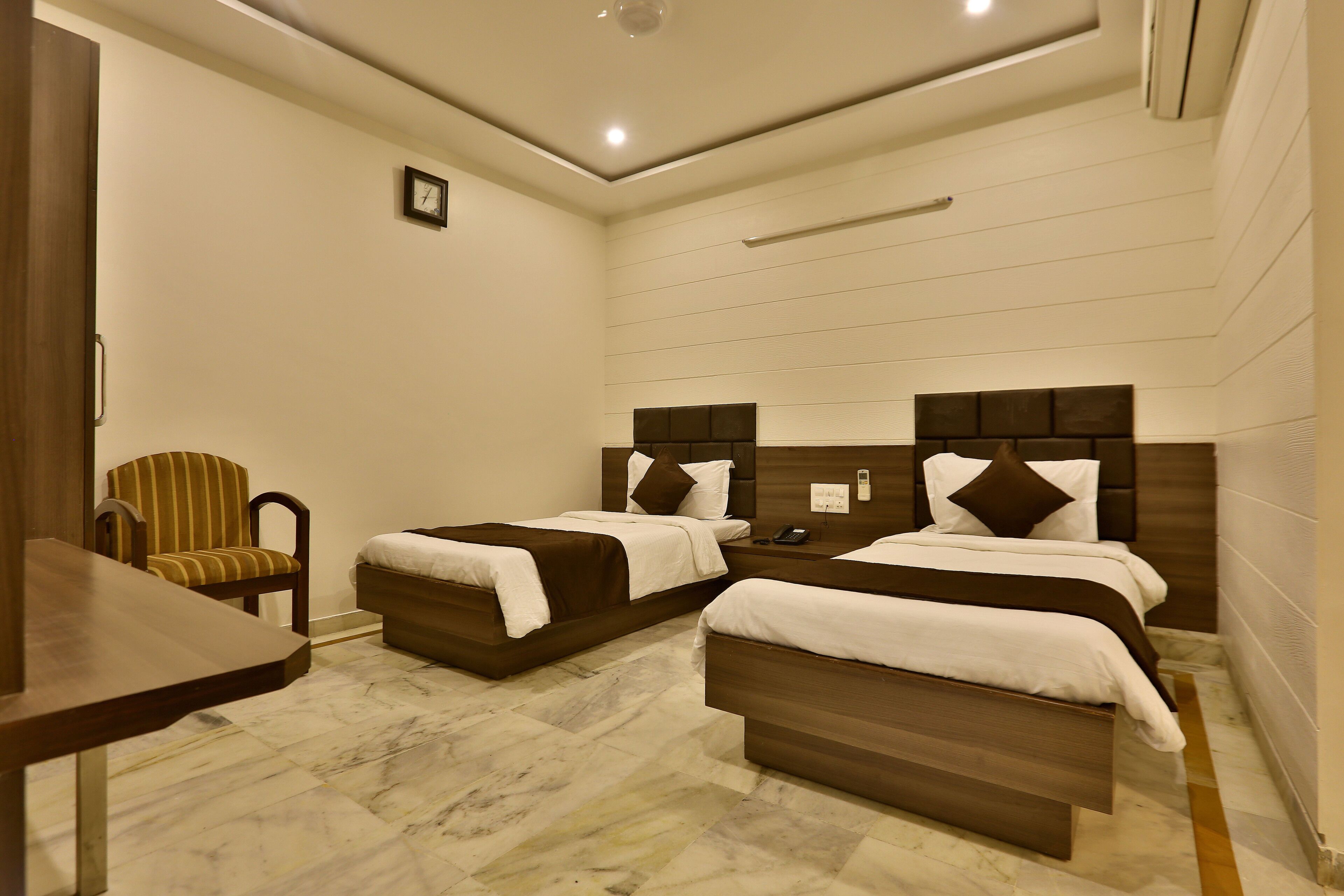 A hotel room with two beds, a wooden chair, a small table, and a clock on the wall.