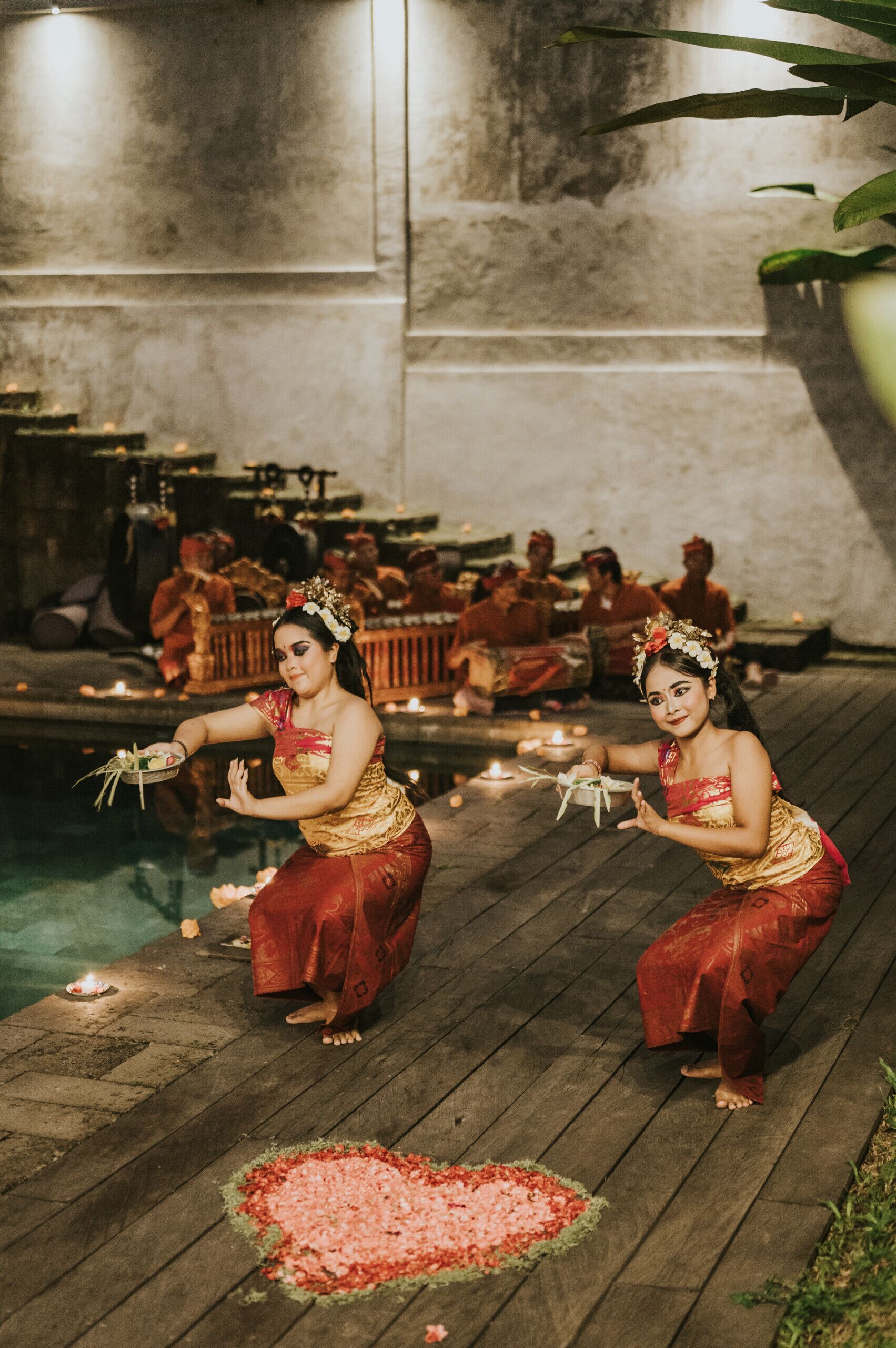 A traditional ceremony with participants in red and gold attire on a wooden platform by a pool, with candles and flowers.