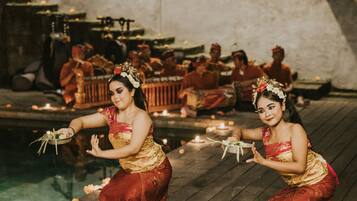 A traditional ceremony with participants in red and gold attire on a wooden platform by a pool, with candles and flowers.