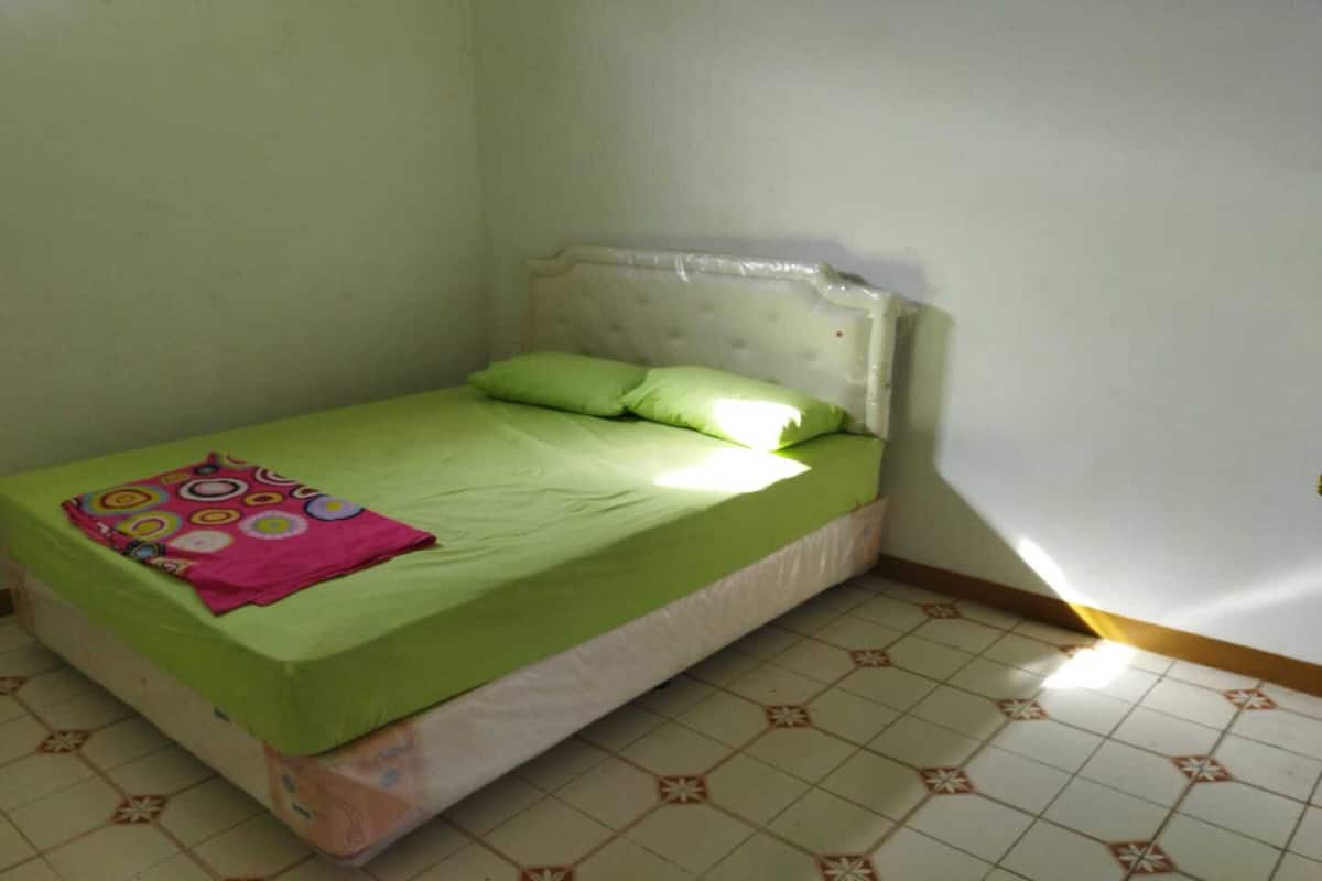 A small, single-bed room with a green bedspread, a pink blanket, and a white headboard. The room has a tiled floor with a simple pattern.