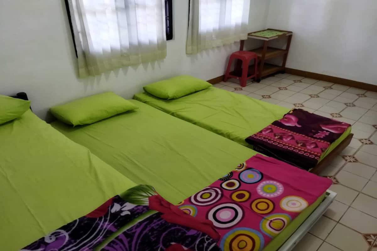 A room with three single beds, each with green bedding. There is a small wooden table with a chair and a window with sheer curtains.