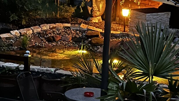 Un espace extérieur aménagé en terrasse, avec un mur en pierre, un petit élément décoratif aquatique et une table ronde entourée de chaises.