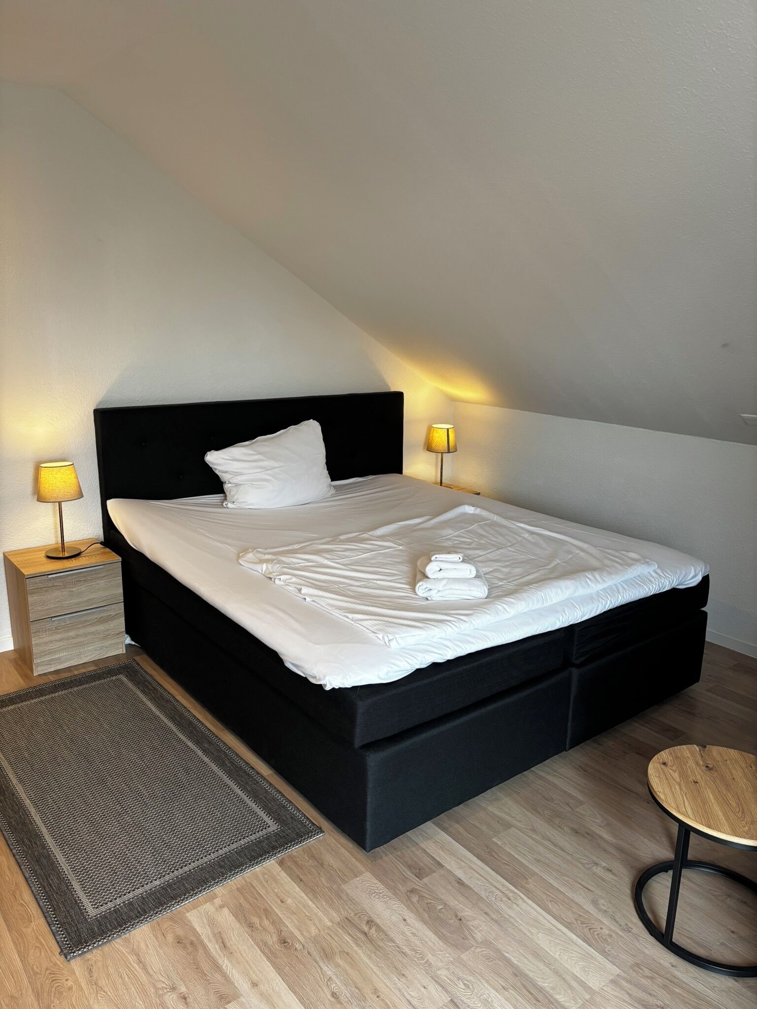 A neatly made bed with white linens and a black headboard in a room with wooden flooring.