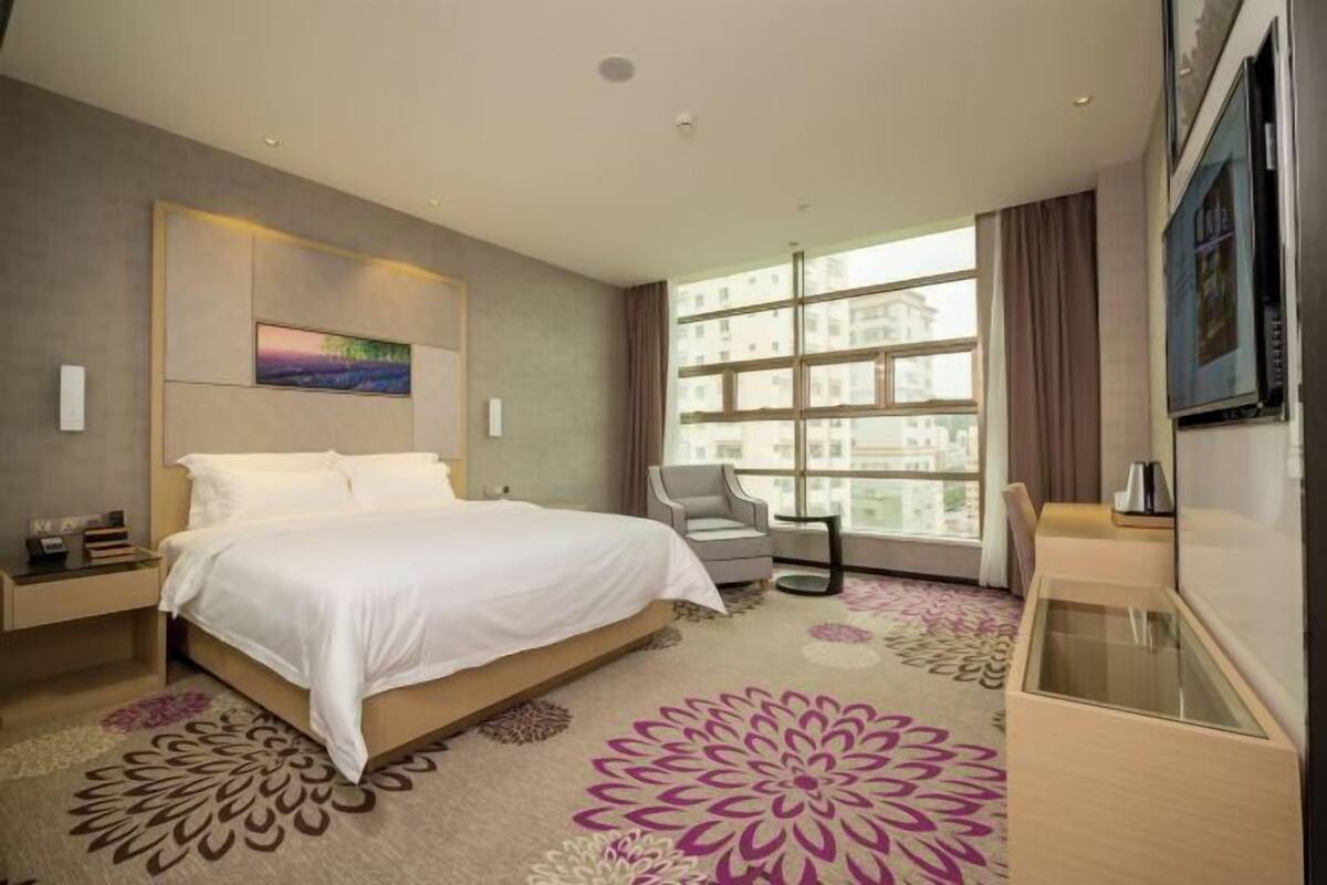 A hotel room with a bed, a chair, a desk, a large window, and a painting on the wall.