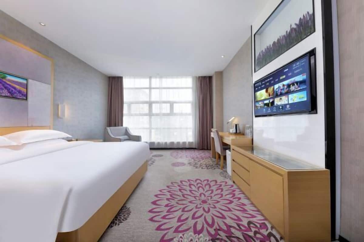 A hotel room with a bed, a desk, a chair, a TV mounted on the wall, and a large window.