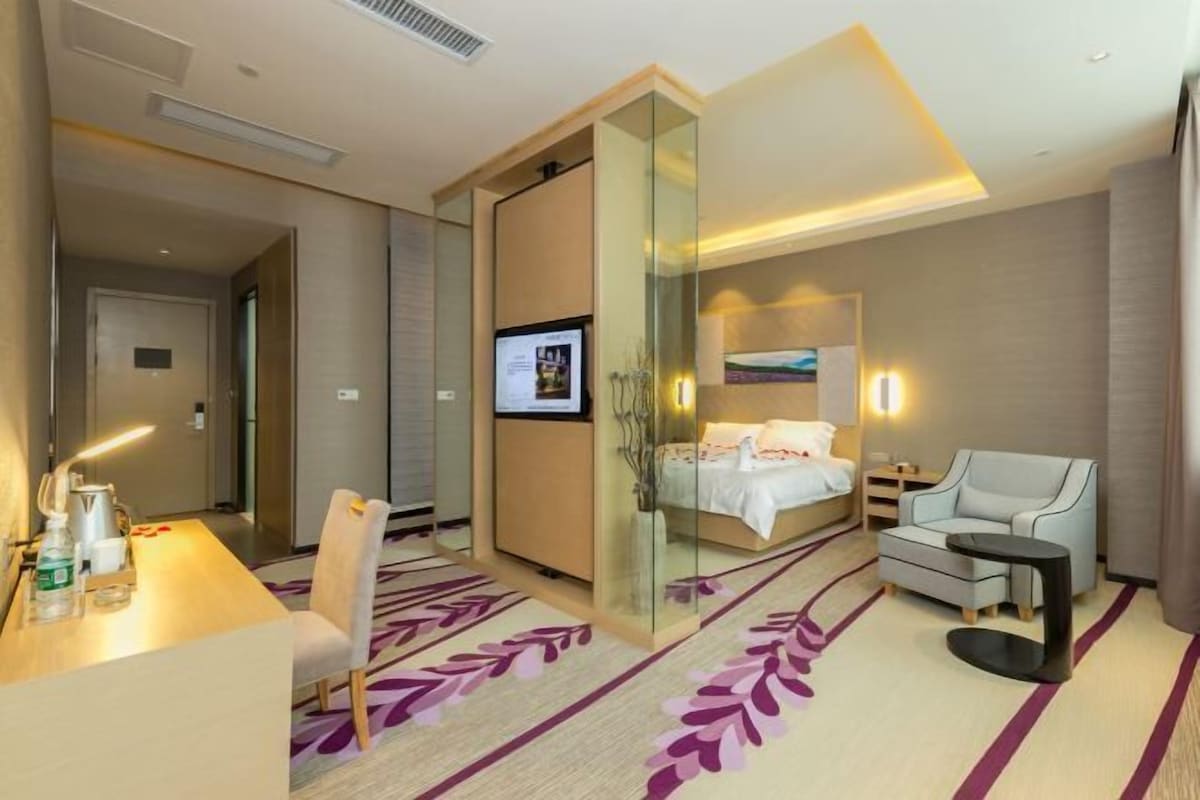 A hotel room with a bed, a desk, a chair, a small table, and a glass partition.