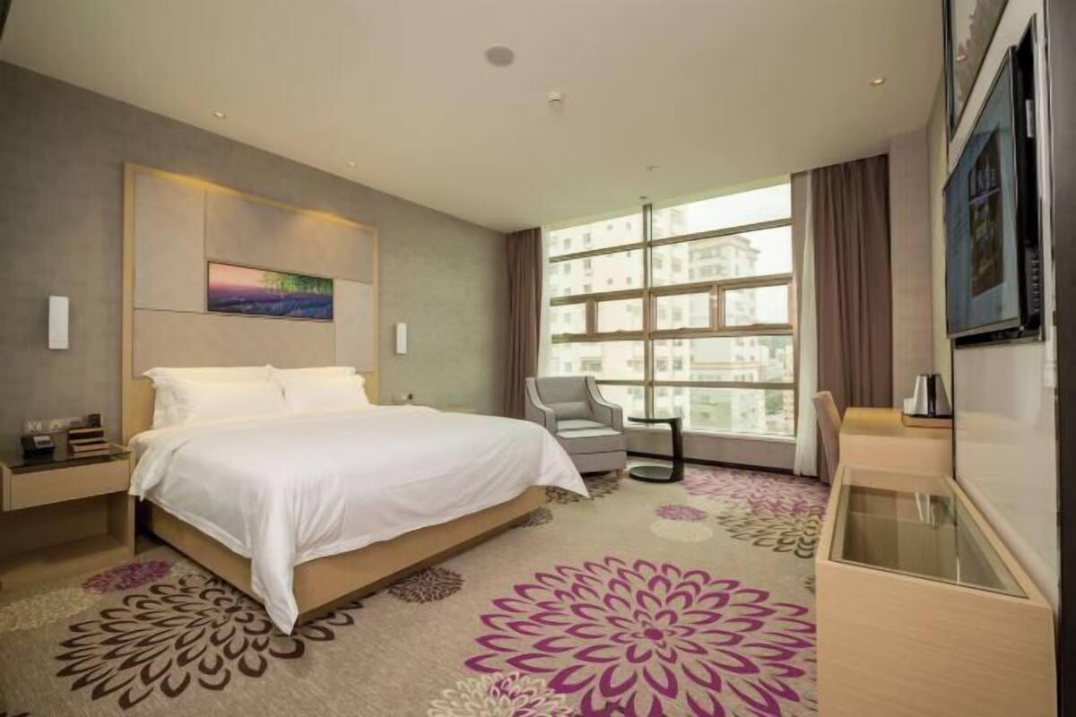 A hotel room with a bed, a chair, a desk, a TV, and a large window with a city view.