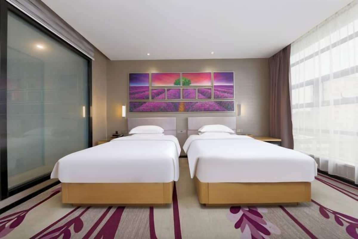 A hotel room with two beds, a large window with curtains, a wall-mounted artwork, and a patterned carpet.