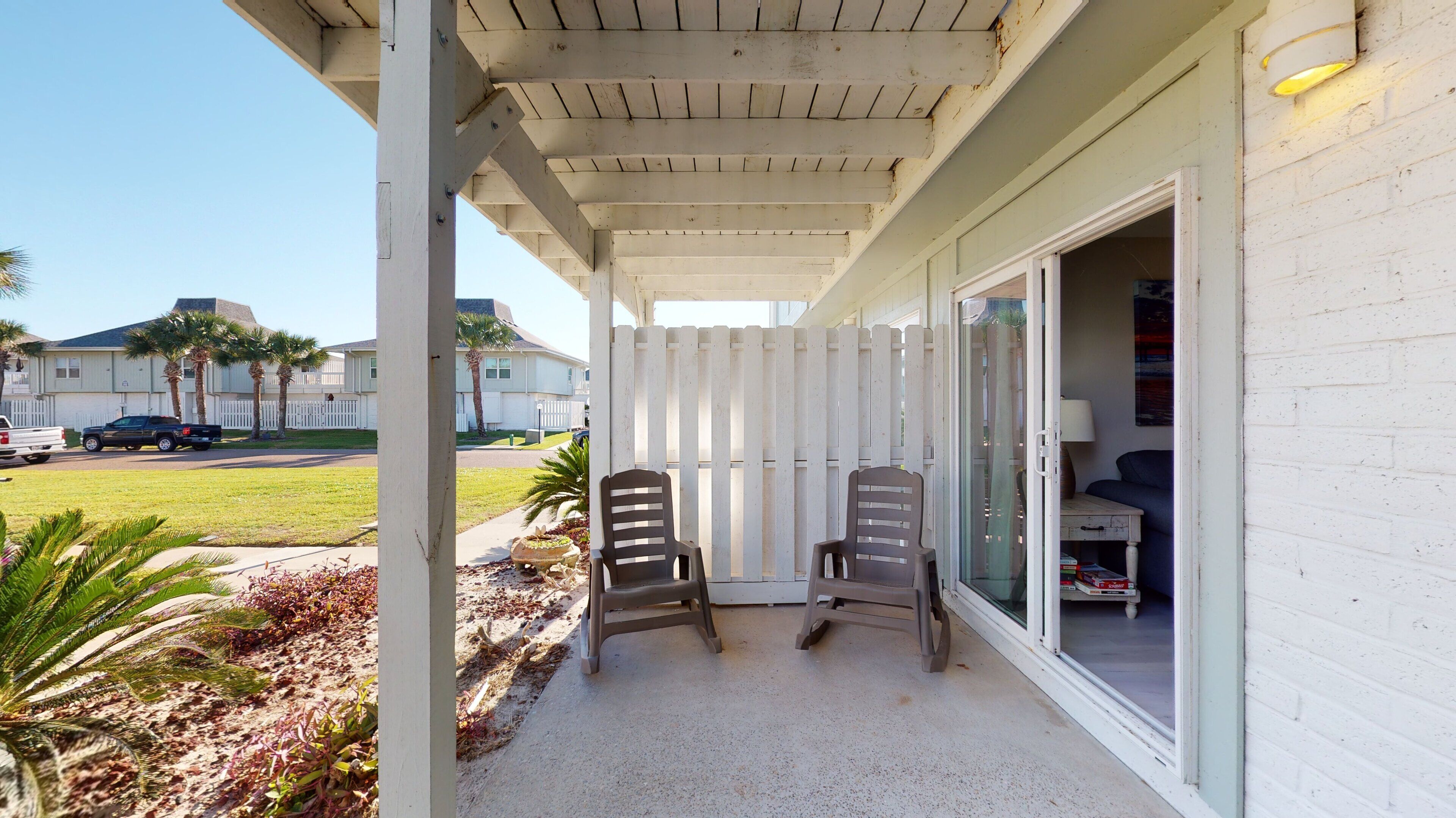 A covered patio area with two chairs, a glass door leading inside, and a view of a residential street with houses and parked cars.