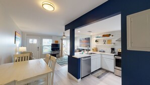 A modern kitchen with white cabinets, a built-in oven, and a coffee maker. There is a dining area with a table and chairs, and a TV in the background. - Ground Floor Condo, Boardwalk to Beach, Shared Pool and Grill (Port Aransas)