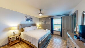 Un dormitorio con cama, lámparas de noche, ventilador de techo, ventana con cortinas, puerta y televisor sobre un aparador de madera.