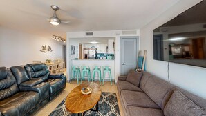 Een woonkamer met een zwarte leren bank, een bruine bank en een ronde houten salontafel. Op de achtergrond staan drie turquoise barkrukken en een keuken.