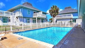 A swimming pool with a white fence, a palm tree, and a two-story building with balconies. - Ground Floor Condo, Shared Pool, Grills and Boardwalk (Port Aransas)