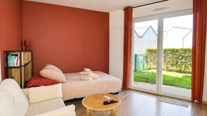 A bedroom with a bed, a sofa, a small round table, a bookshelf, and a sliding glass door to the outside. - La Patoune - Garden/50m from the beach/Wifi/Parking (Courseulles-sur-Mer)
