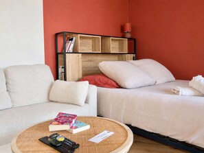 A room with a sofa, a bed, a small round table with books and a remote control, and a wooden bookshelf with books and a lamp.