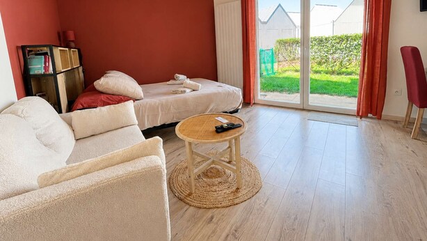 A modern living room with a sofa, a round coffee table, and a bed with a view of the outdoors. - La Patoune - Garden/50m from the beach/Wifi/Parking (Courseulles-sur-Mer)