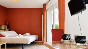 A hotel room with a bed, a table with a red chair, a television, and a window with curtains. - La Patoune - Garden/50m from the beach/Wifi/Parking (Courseulles-sur-Mer)