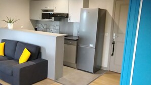 A compact living space with a grey sofa, a small white table, and a kitchen area featuring a microwave, stove, and refrigerator. - LUMINOUS COMFORT CALM (Rouen)