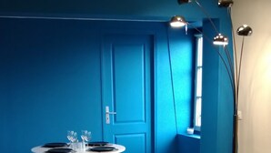 A modern living room with a blue accent wall, a round white table with black plates and glasses, a black sofa with yellow cushions, and a floor lamp. - LUMINOUS COMFORT CALM (Rouen)