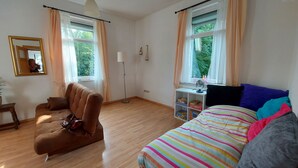 A bedroom with a wooden floor, a brown sofa, a bed with colorful striped bedding, a small white desk with a fan, and a window with curtains. - Apartment at Levinscherpark, 3km from downtown (Göttingen)