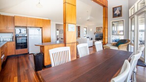 A modern kitchen with wooden cabinets, a dining area with a large wooden table, and a view of the living room with a sofa and chairs. - 3 Buoys - Set high with sweeping ocean views to Moreton Island and Cylinder Beach. (Point Lookout)
