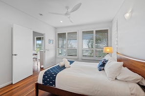 Une chambre à coucher comprenant un lit, un ventilateur de plafond, une fenêtre avec des stores et une porte donnant sur une salle de bain.