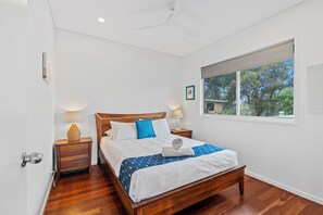 A bedroom with a wooden bed, two bedside tables with lamps, a window with a view of trees, and a wall-mounted picture.