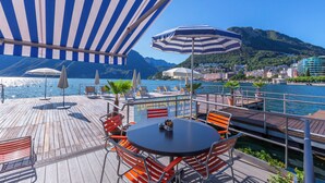 A wooden deck with outdoor seating, a table, and umbrellas overlooking a body of water with mountains in the background. - Boutique Apartment 6 (Lugano)