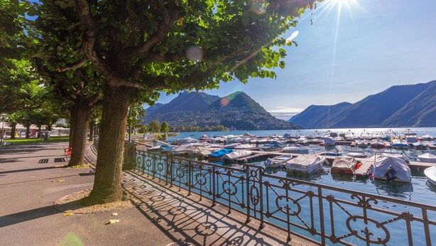 A marina with boats docked, a tree-lined walkway, and mountains in the background. - Boutique Apartment 6 (Lugano)