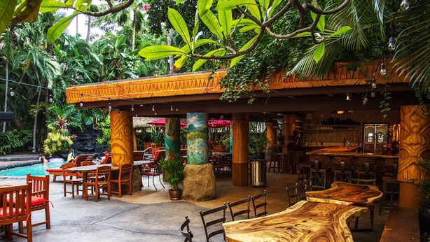 Ein Außenbereich eines Restaurants mit Holztischen und -stühlen, einem Pool und tropischen Pflanzen.