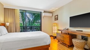 A hotel room with a bed, a desk, a chair, a television, and a view of palm trees through a large window.
